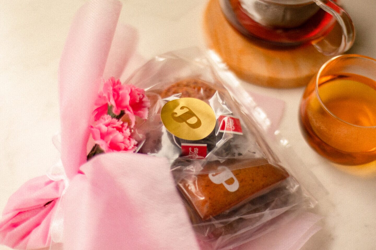 Pink gift wrap with a small bouquet of pink carnations and a clear bag containing cookies, next to two cups of tea.
